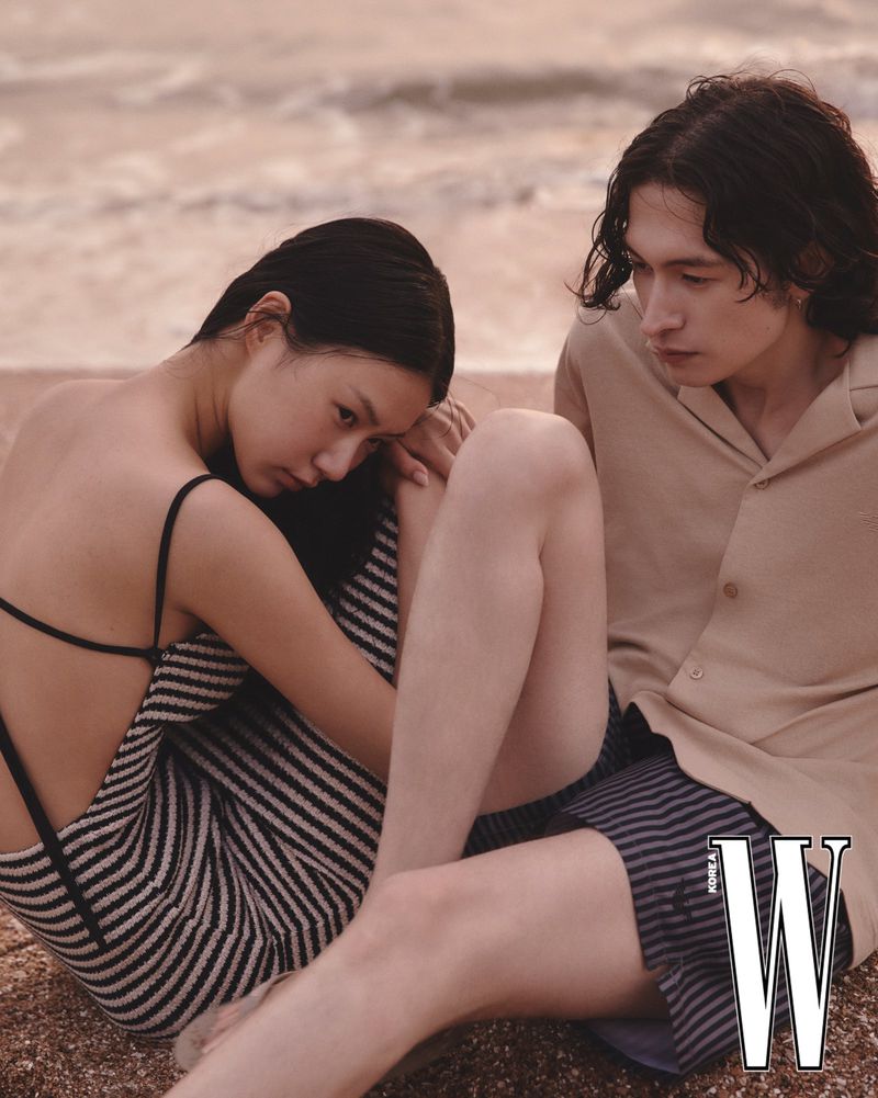 A fashionable couple poses on a sandy beach with ocean waves, the woman in a striped dress and the man in a light shirt and dark shorts.
