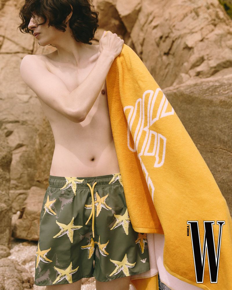 Young adult male poses outdoors on a rocky beach with swim shorts and a yellow towel in a fashion editorial. Magazine logo visible.