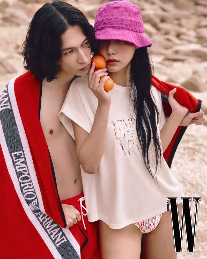 A couple poses outdoors. The woman in a pink hat holds oranges. The man wears a beach towel with an Armani logo. Magazine advertisement.