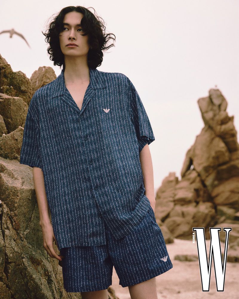 A long-haired male model poses in a navy blue striped short-sleeve shirt and shorts on a rocky beach, in front of a W Magazine logo.