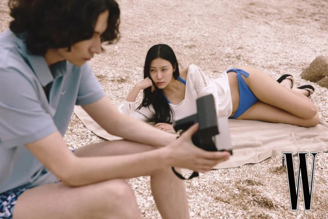 Fashion editorial shot on a beach with a photographer and a woman in a blue bikini and white cover-up posing in natural light.