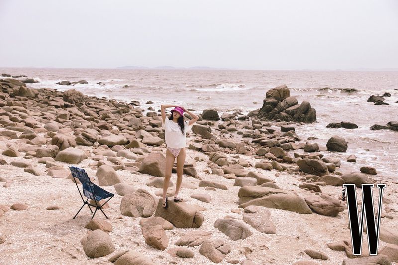 Model Hojin Lee stands on a rocky beach wearing a white t-shirt, pink bucket hat, and swimwear beside a blue folding chair.