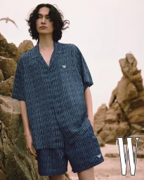 A male model with dark curly hair wearing a blue patterned short sleeve shirt and shorts posing against rocky beach formations