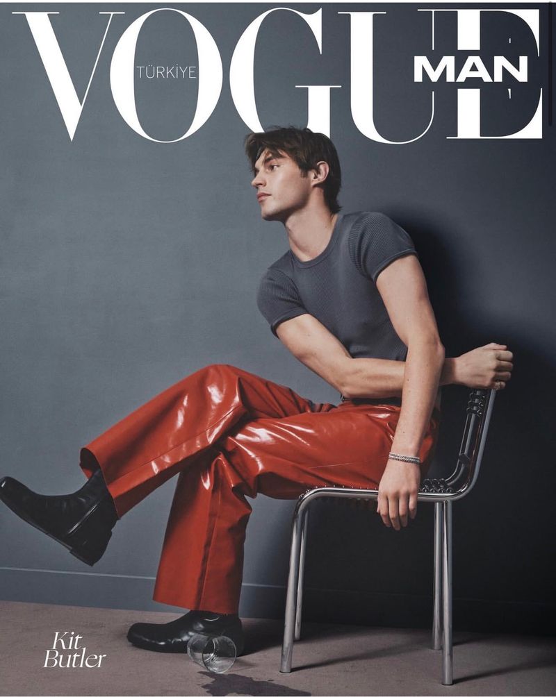 Model Kit Butler seated sideways on a chrome chair in glossy red trousers and a grey t-shirt against a dark backdrop.