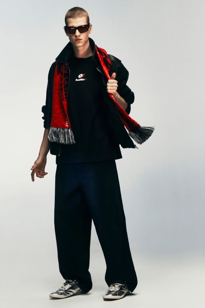 Male model in black jacket and trousers wearing sunglasses and a red scarf against a white background