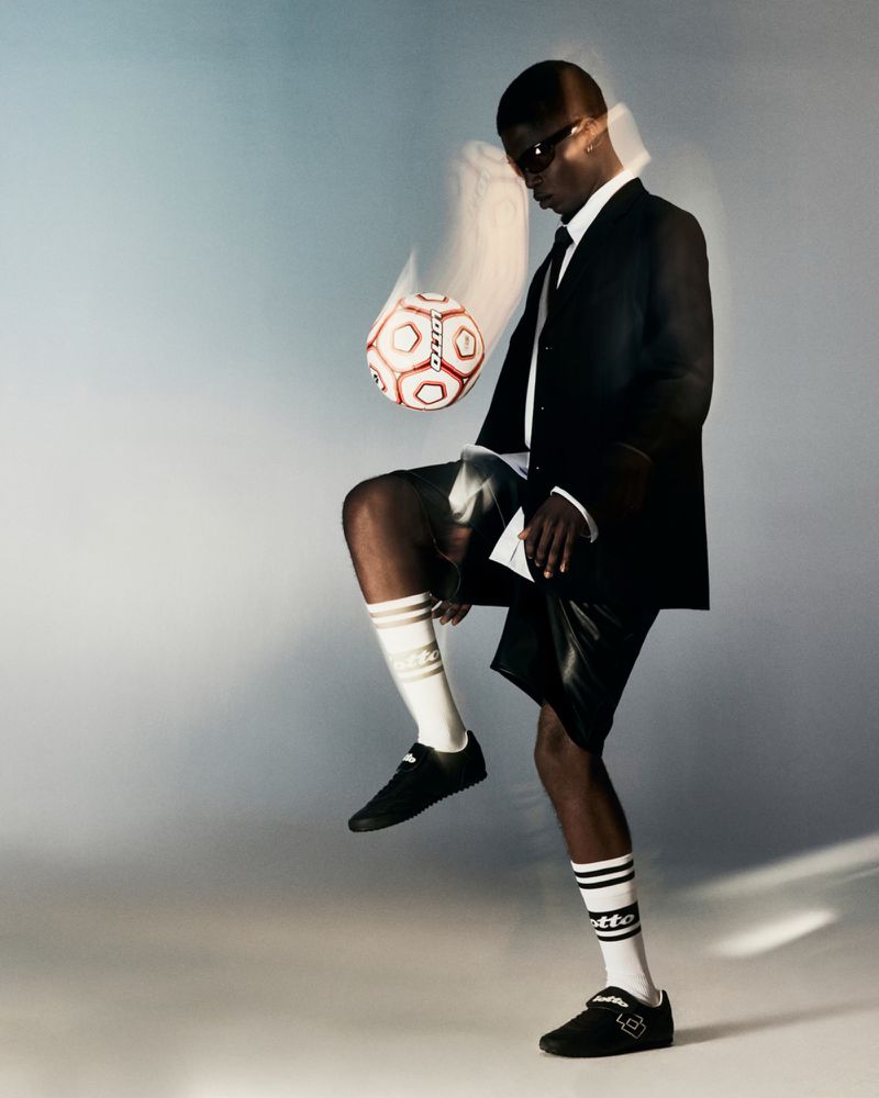 Model Jeremiah Berko Fourdjour balances a soccer ball on his knee wearing a black suit jacket, athletic shorts, and branded socks.