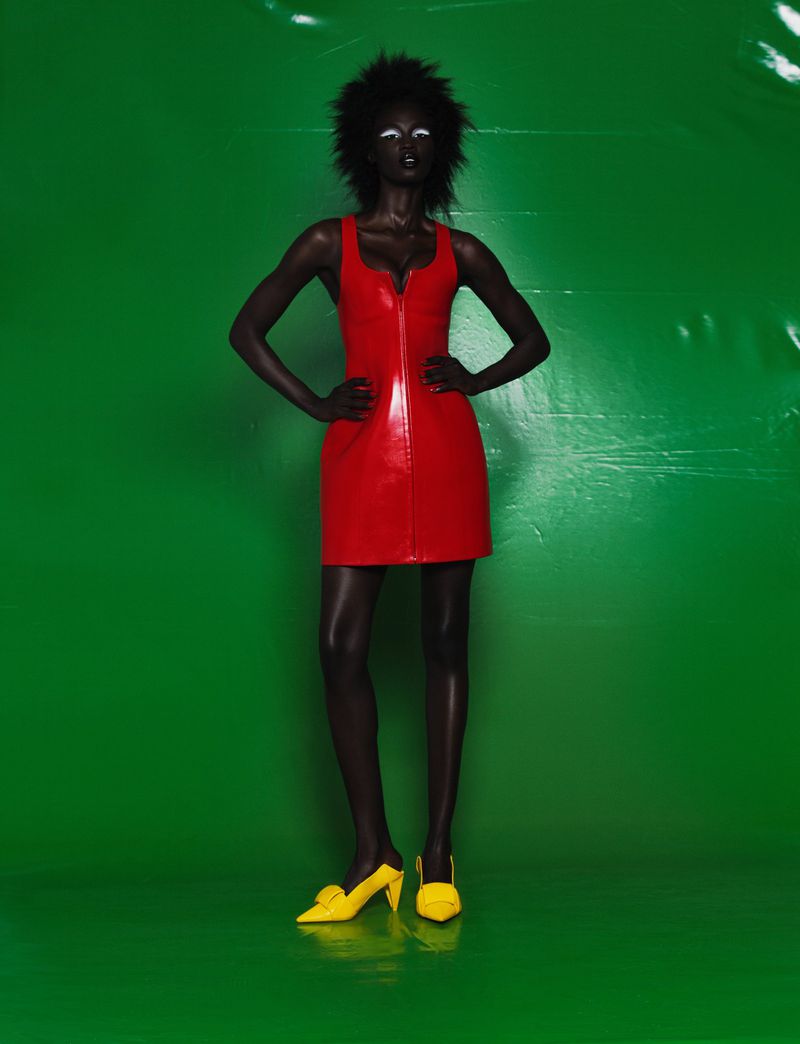 Model Sokhna Cisse wearing a red sleeveless mini dress and yellow pointed heels against a solid green background.