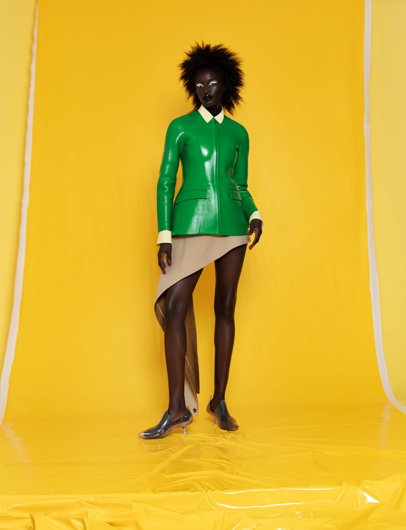 Model wearing a glossy green jacket and asymmetric tan skirt posing against a bright yellow seamless paper background.