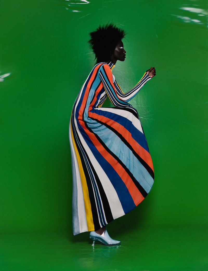 Dark-skinned model poses in a colorful striped dress and light blue heels against a bright green studio backdrop, fashion photoshoot.