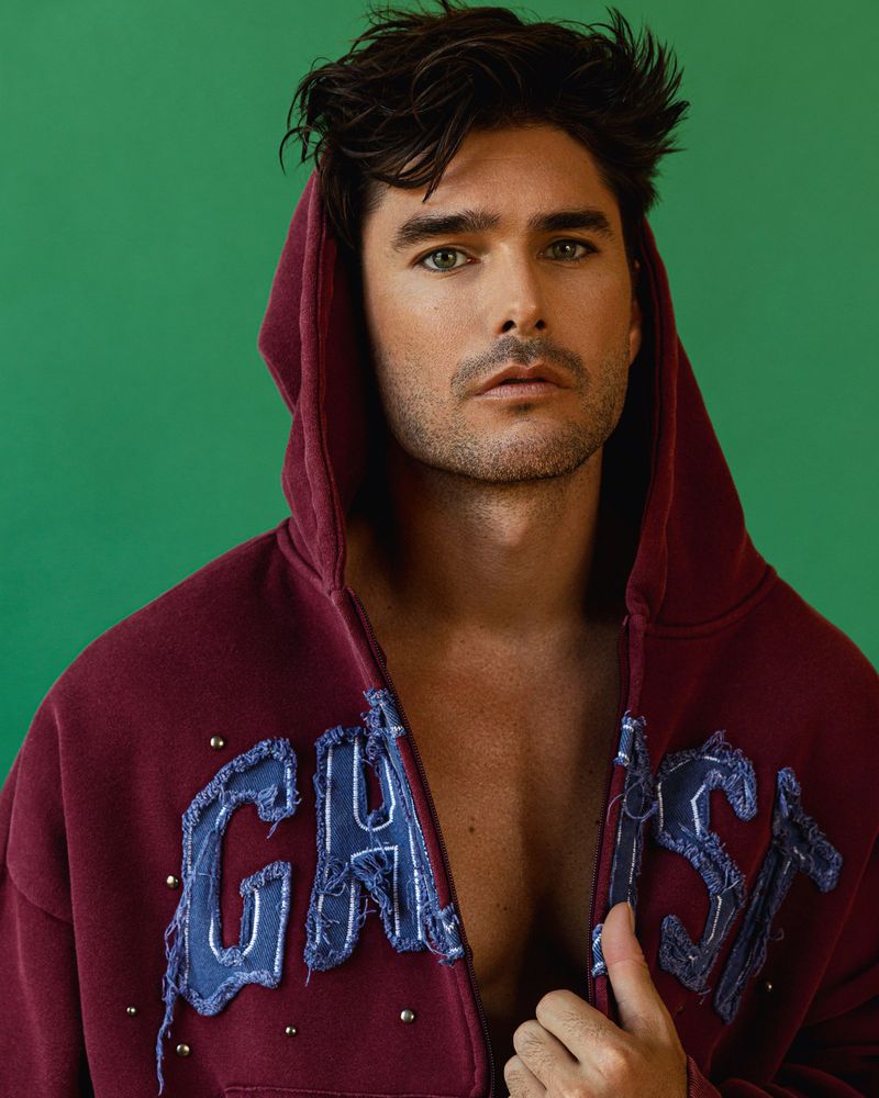 Model Charlie Matthews wearing an unzipped burgundy hoodie with hood raised against a green background.