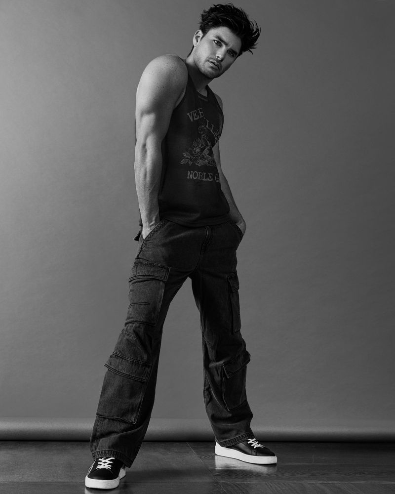 Model Charlie Matthews posing in a dark graphic tank top and black wide-leg cargo pants against a studio backdrop.
