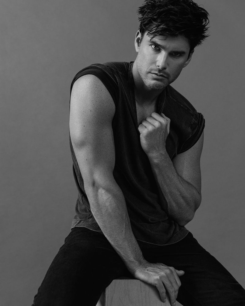 Black and white portrait of Charlie Matthews seated in a sleeveless shirt against a grey studio backdrop.