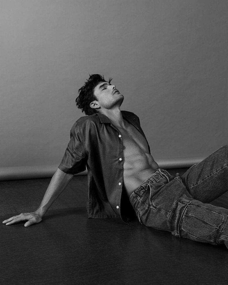 Model Charlie Matthews leaning back on a dark studio floor wearing an unbuttoned shirt and denim jeans