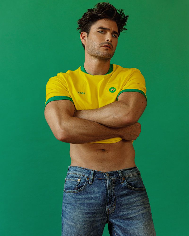 Model Charlie Matthews wearing a yellow ringer t-shirt and blue jeans with arms crossed against a green background.
