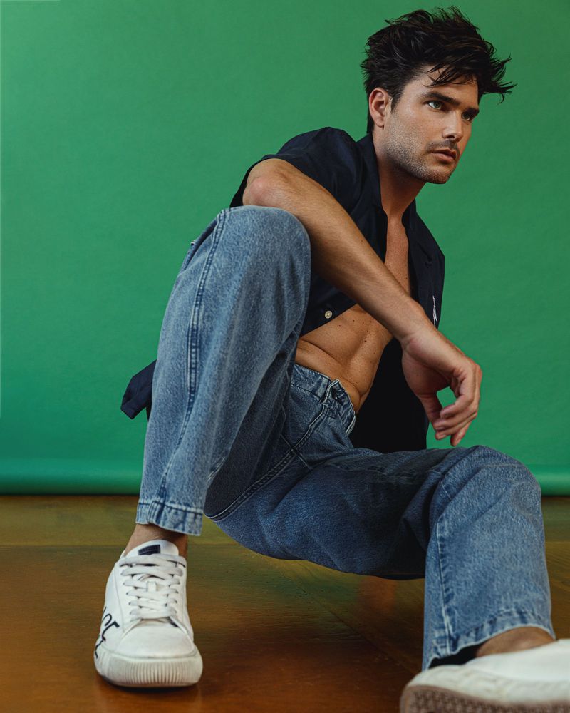 Male model Charlie Matthews seated wearing unbuttoned dark shirt, blue jeans and white sneakers against a vibrant green backdrop.