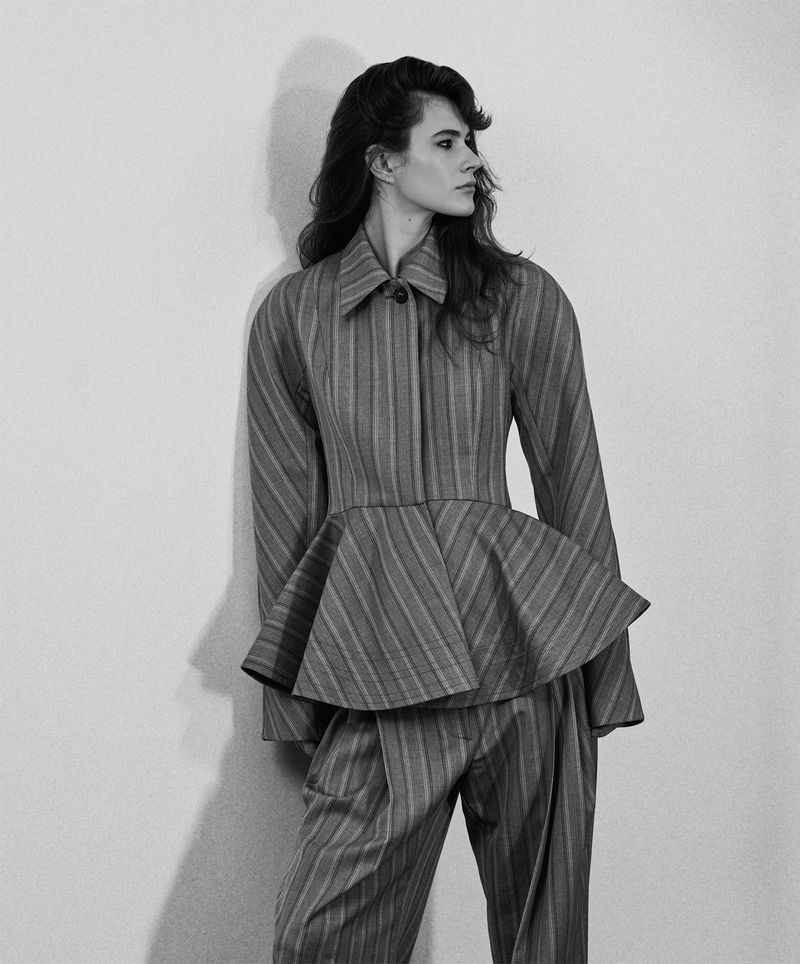 Model in a striped peplum jacket and matching trousers leaning against a light wall in a monochrome fashion portrait.