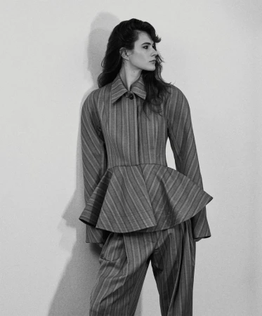 Model in a striped peplum jacket and matching trousers leaning against a light wall in a monochrome fashion portrait.