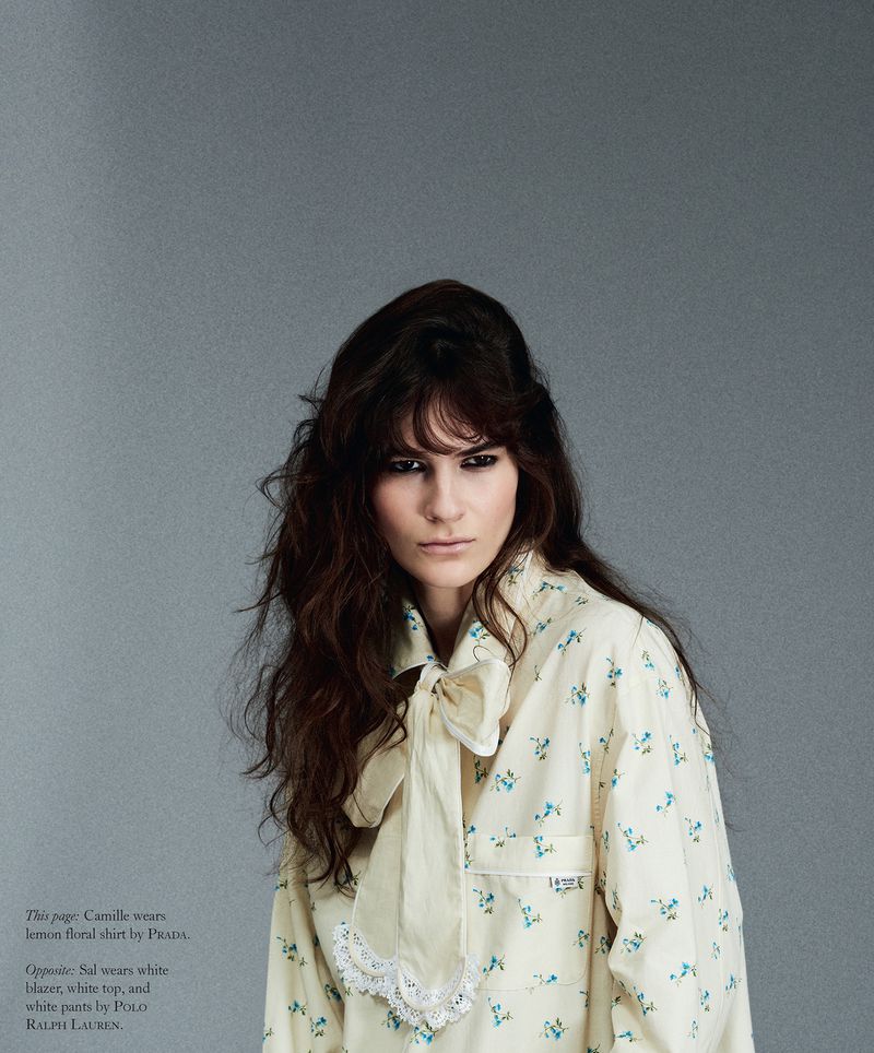 Model with long dark hair wearing a cream floral blouse with a neck bow against a solid grey background.