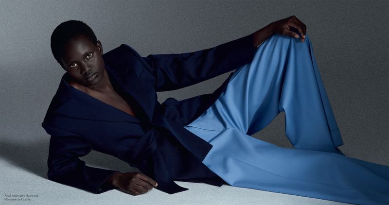 Model Aluel Keror reclining on a textured grey studio floor wearing a dark navy blazer and wide-leg light blue trousers.