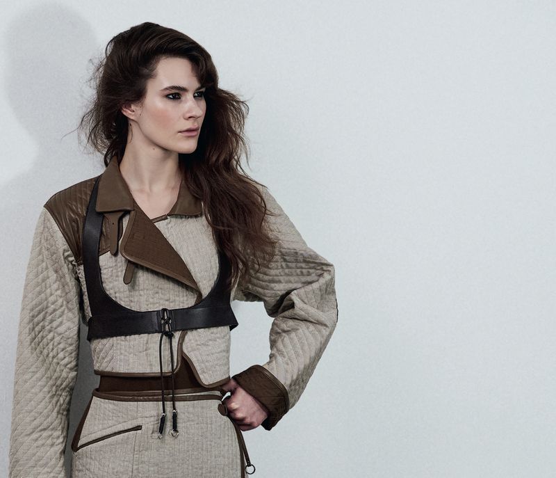 Model wearing a quilted beige jacket and dark leather harness against a white seamless background