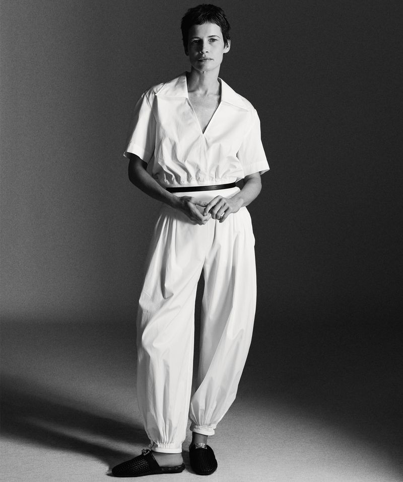 Model with short dark hair wearing a white jumpsuit standing against a grey studio backdrop.