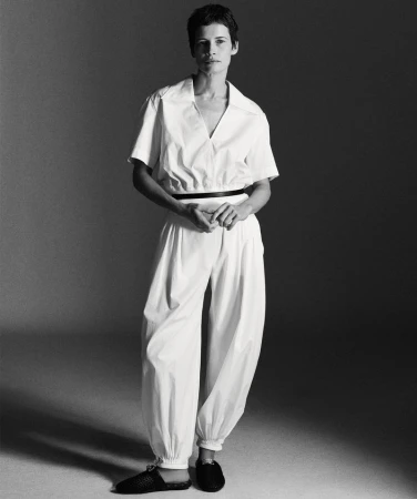 Model with short dark hair wearing a white jumpsuit standing against a grey studio backdrop.