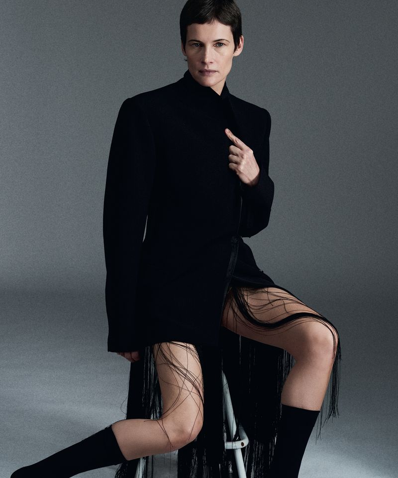 Model with short dark hair seated on a stool wearing a black jacket and fringed garment against a grey background.