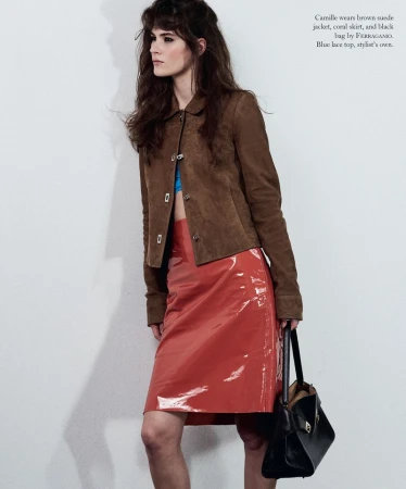 Model Camille Desjardins posing in a brown suede jacket, glossy coral skirt, and holding a black handbag against a white studio backdrop.