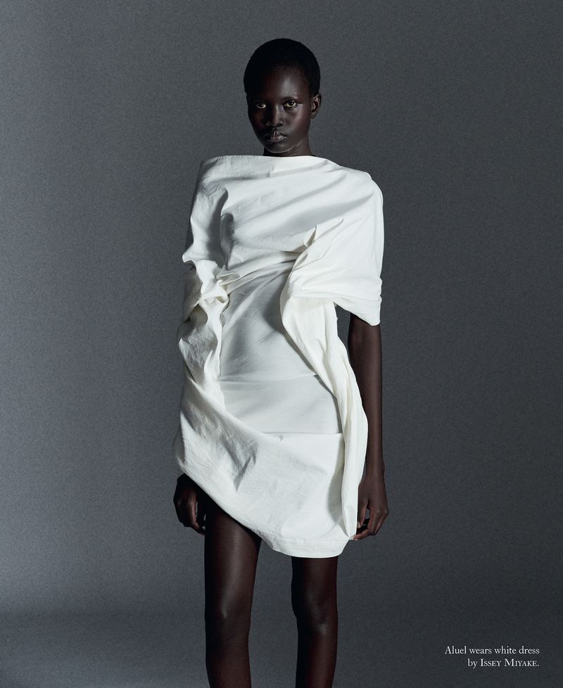 Model Aluel Keror posing in a sculptural draped white mini dress against a smooth grey studio backdrop.