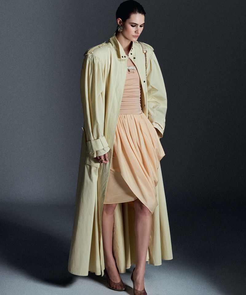 Model wearing a pale yellow long coat and peach dress against a grey studio seamless backdrop