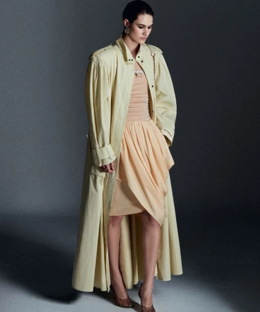 Model wearing a pale yellow long coat and peach dress against a grey studio seamless backdrop