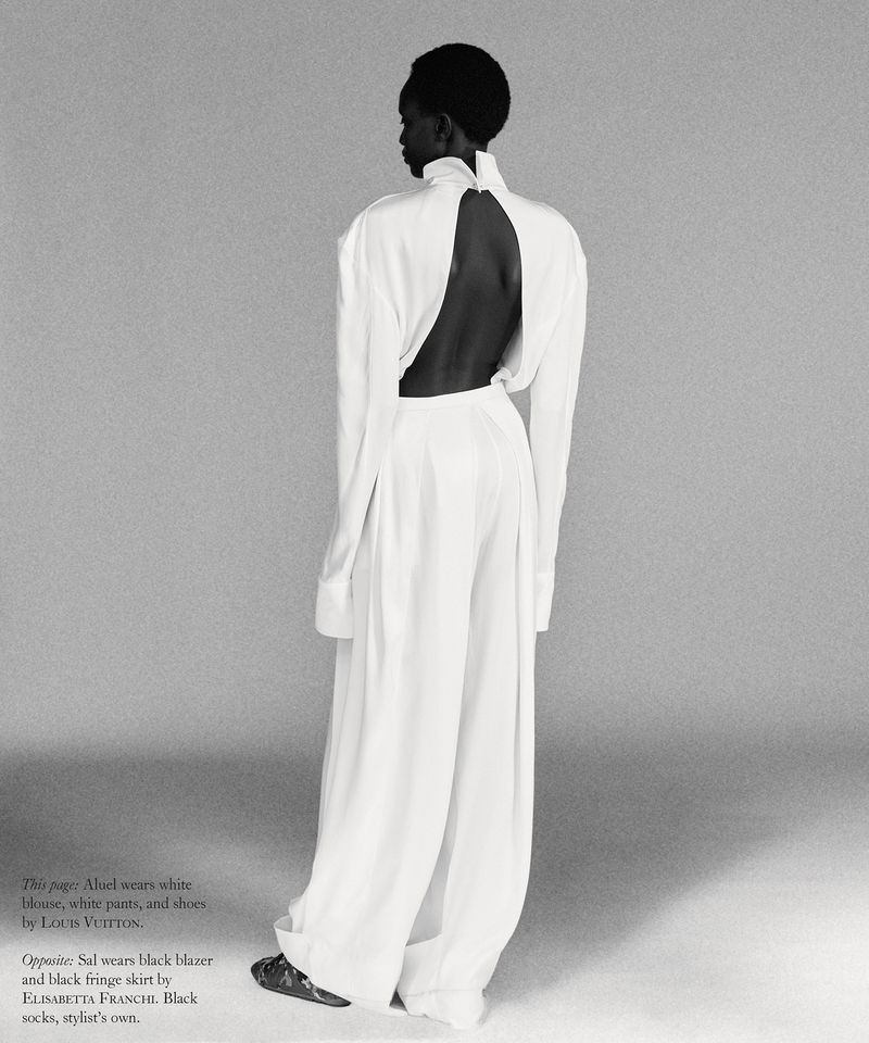 Model standing with back turned wearing a white high-neck blouse and wide-leg trousers against a grey seamless studio backdrop.
