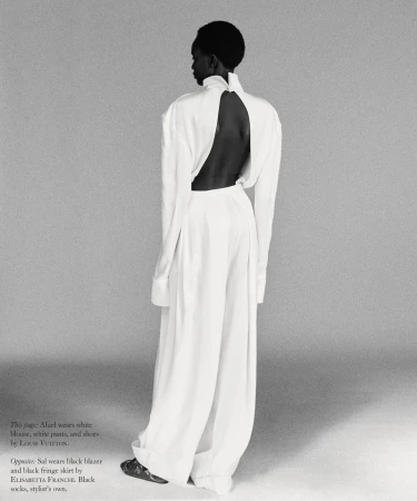 Model standing with back turned wearing a white high-neck blouse and wide-leg trousers against a grey seamless studio backdrop.