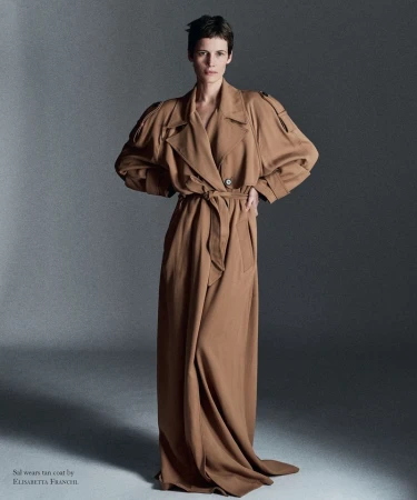 Model with short dark hair standing in an oversized, floor-length tan trench coat tied at the waist against a grey studio backdrop.