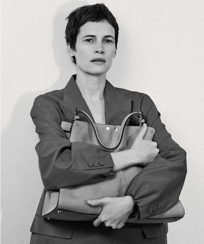 Model with short dark hair in an oversized blazer clutching a large structured tote bag to her chest against a neutral backdrop.