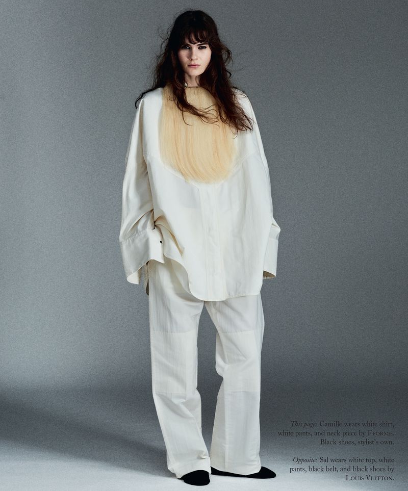 Model Camille Desjardins standing in an oversized white shirt, white trousers, and a pale yellow fringe bib on a grey background.