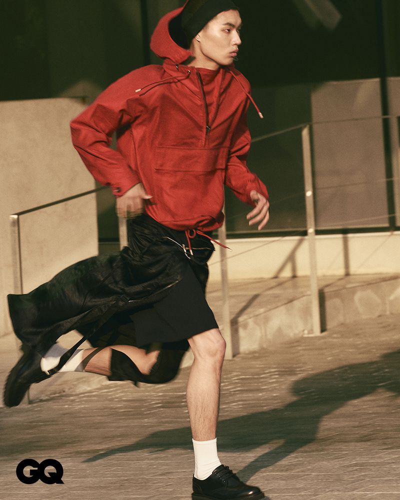 Asian male fashion model in motion wearing red hooded jacket, black shorts, black sneakers, and white socks on an urban street.