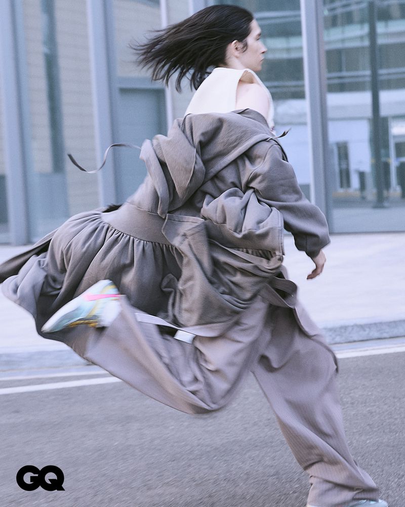 Fashion editorial shot of a person in an oversized coat running in an urban environment, with a modern building and grey pavement.
