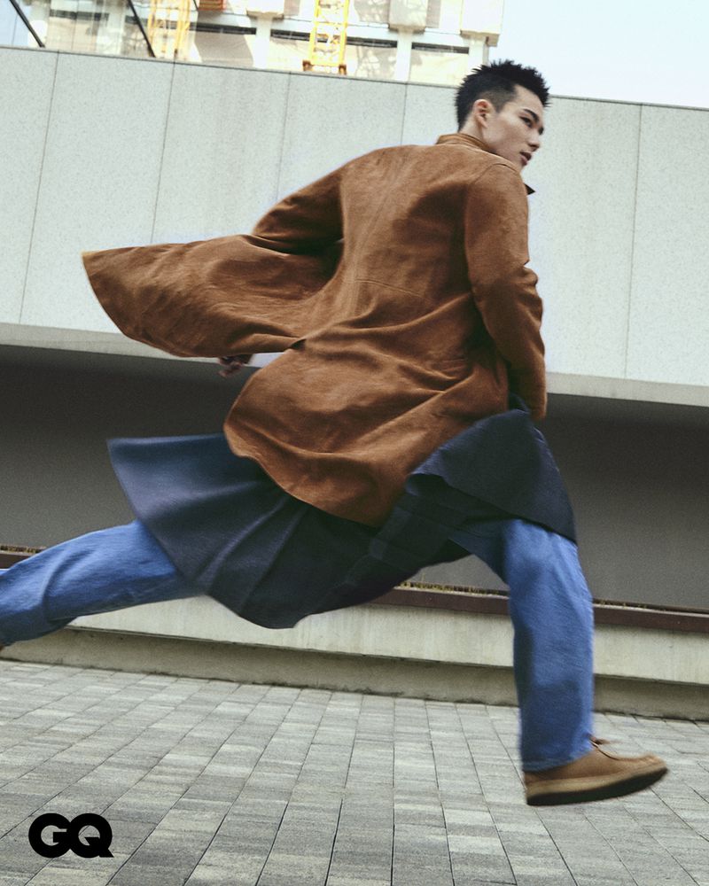 Asian male model in motion wearing a tan overcoat, blue jeans, and tan shoes on a city street with modern architecture in the background.