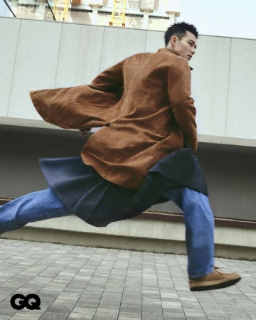 Asian male model in motion wearing a tan overcoat, blue jeans, and tan shoes on a city street with modern architecture in the background.