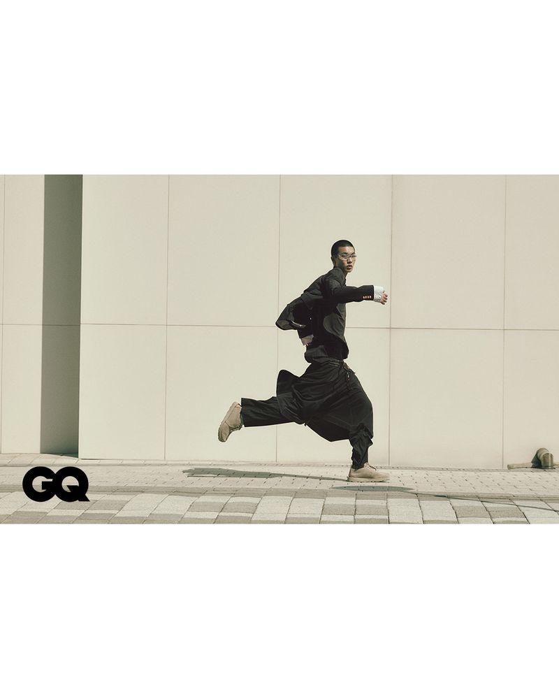 Asian man runs in a black jacket and pants near a modern building. He is in motion on a brick sidewalk with a logo on the side.