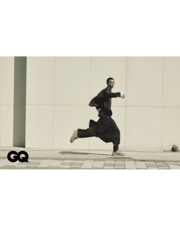 Asian man runs in a black jacket and pants near a modern building. He is in motion on a brick sidewalk with a logo on the side.