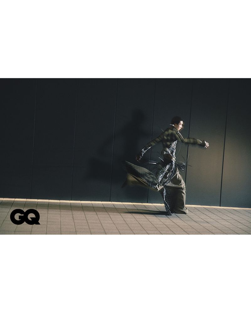 Fashion editorial shot of an adult male model wearing a sculptural green and grey coat, posing in a running stance on a tile floor.