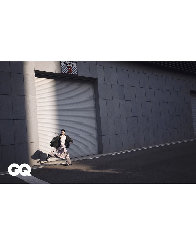 Fashion editorial image shows a male model in a patterned outfit running in front of a gray wall with industrial doors and signage.