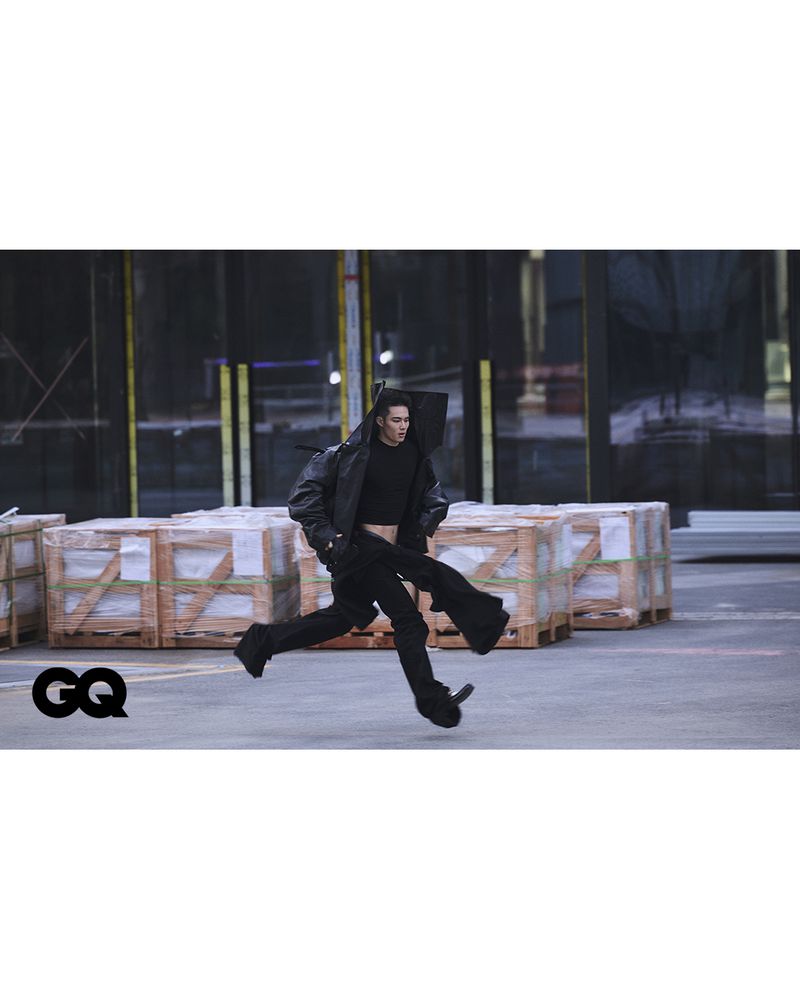 A person runs on a city street in an open black leather jacket, pants, and a crop top with GQ logo and industrial background.
