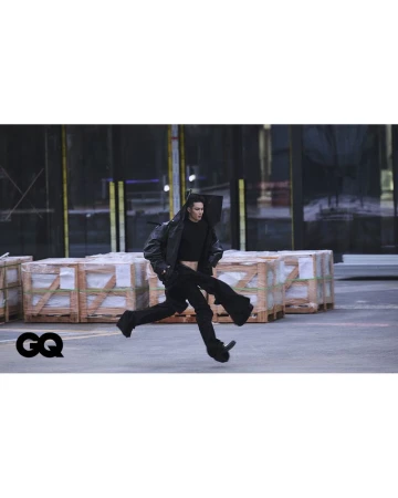 A person runs on a city street in an open black leather jacket, pants, and a crop top with GQ logo and industrial background.