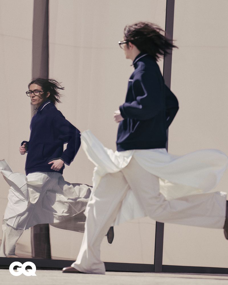 A man in a dark jacket and white pants runs past a reflective wall, his reflection also running, captured in an urban setting with GQ logo.
