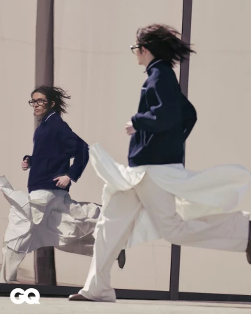 A man in a dark jacket and white pants runs past a reflective wall, his reflection also running, captured in an urban setting with GQ logo.
