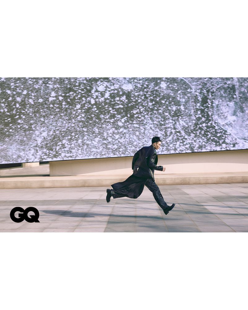 Fashion editorial shot of a man in black running outdoors in a long jacket and patterned pants, with a concrete and tile background.