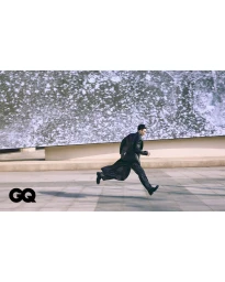 East Asian male model leaping mid-air across a paved plaza wearing a long black coat for GQ Korea.